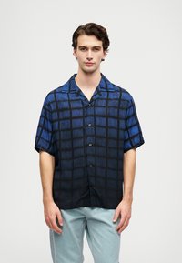 Ung mann som har på seg en kortermet skjorte med blå og svart rutemønster og lyseblå jeans, stående mot en ensfarget bakgrunn.