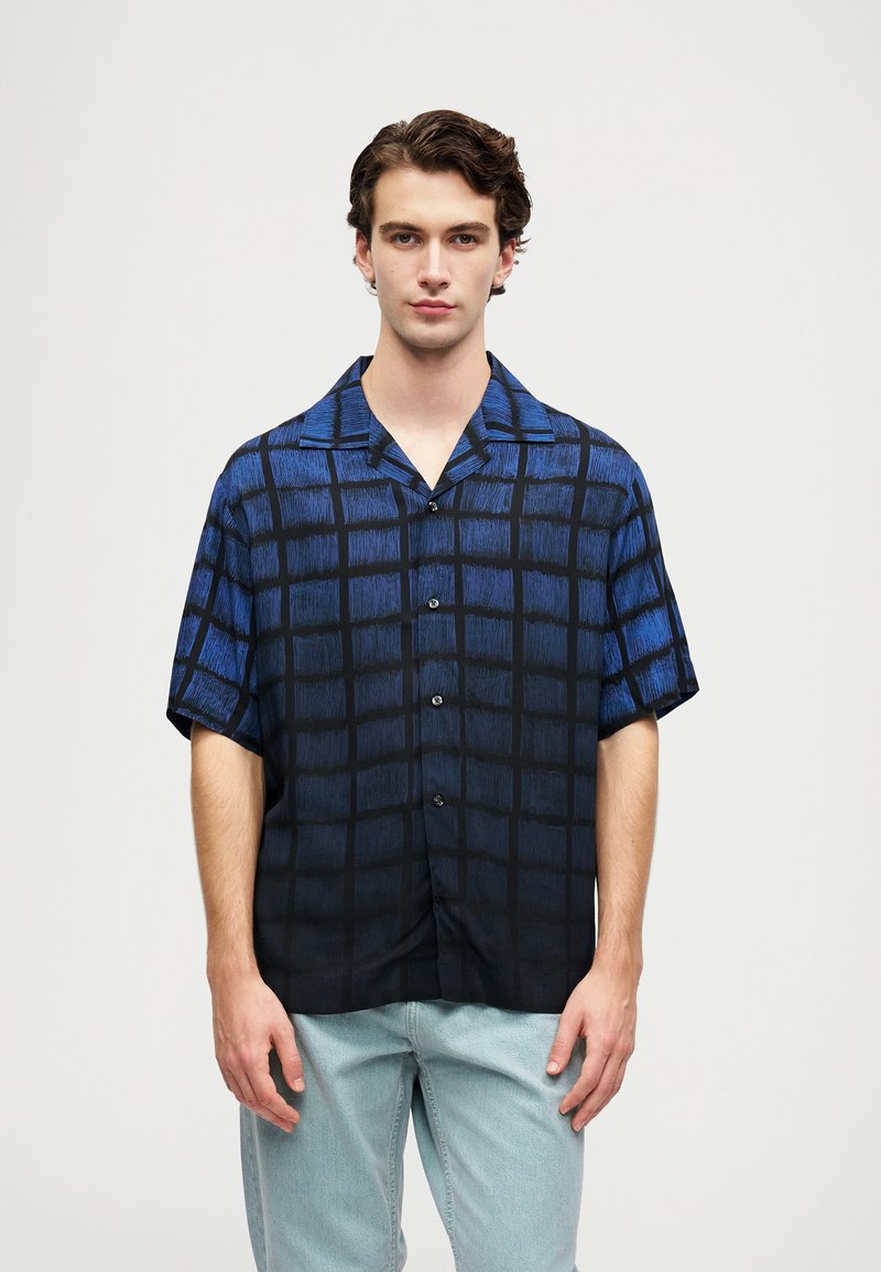 Ung mann som har på seg en kortermet skjorte med blå og svart rutemønster og lyseblå jeans, stående mot en ensfarget bakgrunn.