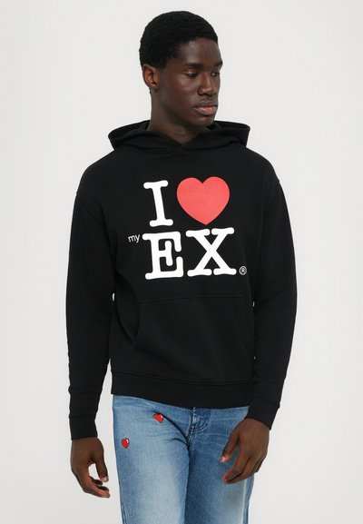 Felpa con cappuccio nera con grafica bianca e rossa che recita "I ♥ my EX." Tasca frontale a marsupio. Indossata con jeans blu con accenti a forma di cuore rossi.