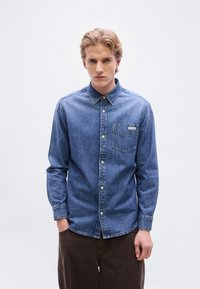 Πουκάμισο από τζιν με κουμπιά σε μεσαίο μπλε, μακριά μανίκια, γιακά, μία τσέπη στο στήθος και μικρή ετικέτα με λογότυπο. Φορεμένο με καφέ παντελόνι.