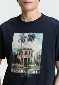 Tums zils kokvilnas T-krekls, kura priekšpusē ir grafika ar vēsturisku ēku un palmu kokiem, ar uzrakstu "HAVANA TIMES" un zīmola nosaukumu zem tās.