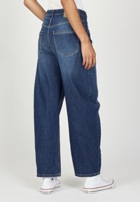 Weite blaue Denim-Jeans mit mittlerem Waschgang, Fünf-Taschen-Design und kontrastierenden Nähten, kombiniert mit weißen High-Top-Sneakern.
