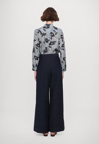 Modrá košile s vzorem a tmavé kalhoty. Košile má květinové vzory a geometrické tvary. Kalhoty jsou široké s manžetou na spodním lemu.