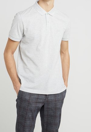 Homme portant un polo à manches courtes gris clair avec col et un pantalon à carreaux gris foncé, les mains dans les poches, debout devant un fond blanc.