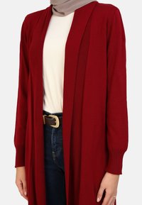 Long, open-front cardigan in deep red knit, featuring ribbed texture and long sleeves, worn over a beige top and dark blue jeans with a belt.