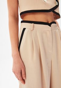Beige zkrácený top a kalhoty s vysokým pasem se černým lemem, zdobené sklady a knoflíky, vyrobené z hladkého, texturovaného materiálu.