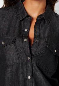Svart denimskjorta med knappar som har krage, två bröstfickor och silverfärgade tryckknappar med en texturerad yta.