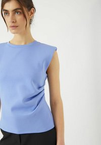 Ljusblå ärmlös topp med rund halsringning, slät textur och subtil draperingsdetalj, ihop med mörka byxor.