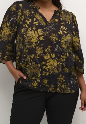 Vrouw draagt zwarte blouse met geel bloemmotief en zwarte broek, met één hand in de zak, tegen een effen achtergrond.