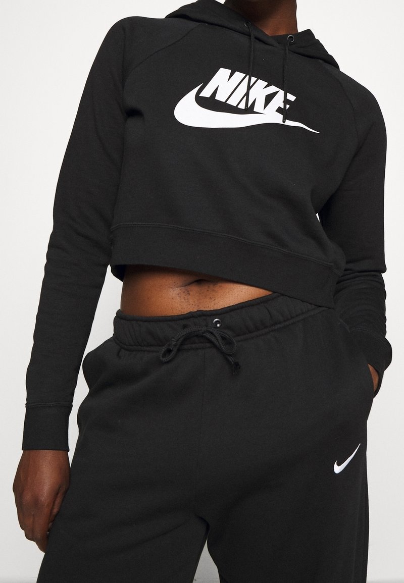 Personne portant un sweat à capuche Nike noir court et un pantalon de survêtement Nike noir, main dans la poche, ventre découvert sur un fond uni.