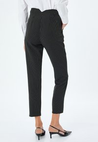 Schwarze Nadelstreifen-Hose mit hoher Taille, schmaler Passform und verkürzter Länge, kombiniert mit schwarzen spitzen Pumps. Glattes Material mit vertikalen Streifen.