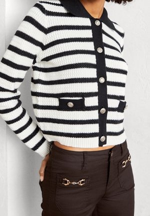 Kvinde iført en sort og hvid horisontalt stribet knap-op cardigan og mørke bukser med guldkædedetaljer på lommerne.