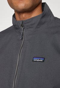 Gray zip-up jacket with a high collar, featuring a Patagonia logo patch on the chest and textured fabric, suitable for outdoor wear.