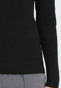 Primo piano di una persona che indossa un maglione a maglia nera con orlo e maniche a costine, abbinato a pantaloni grigi.