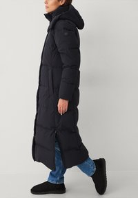 Lang sort pufferjakke med hætte, lynlås foran og elastiske ærmer, med et quiltet design og sidelægger for bedre bevægelsesfrihed.