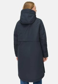 Kvinde iført en lang, mørkeblå vinterfrakke med hætte, set fra ryggen, med lysbrunt hår og blå jeans.