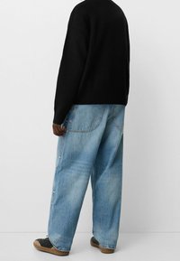 Pull noir en tricot, jean bleu clair ample avec des coutures détaillées, et chaussures noires avec semelles marron. Texture visible sur le pull et le jean.