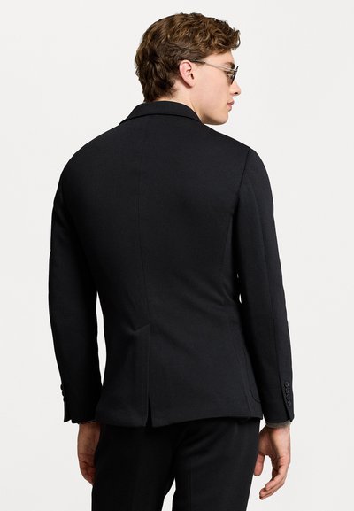 Blazer noir ajusté avec une texture lisse, présentant un empiècement central, des revers crantés et des accents de boutons sur les manches.