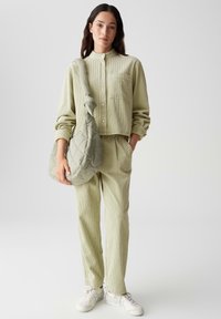 Camicetta corta in un verde chiaro, con texture, colletto alla cinese e taschino frontale, abbinata a pantaloni coordinati e una borsa imbottita verde oliva.