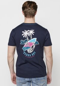 Tumsā zilā kokvilnas t-krekls ar grafiku uz muguras, kurā attēlots skelets sērfingojot, palmu koki, viļņi un teksts "Osaka Bay" zils un rozā krāsā.