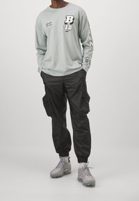 Langärmliges hellblaues Hemd mit schwarzem Print, kombiniert mit schwarzen Cargo-Hosen mit Seitentaschen. Schuhwerk: graue Sportschuhe.