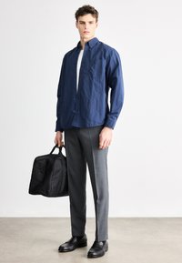Cămașă bleumarin cu nasturi, pantaloni gri, croiți, pantofi negri din piele, ținând o geantă neagră. Textură netedă, design casual și croială mulată.