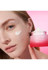 Behållare med en krämig hudvårdsprodukt i en gradient rosa burk märkt "Närande Smältande Mjuk Creme/Mask." Kräm applicerad på kind.