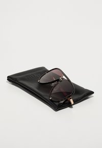 Gafas de sol con lentes oscuros, montura de carey y acentos dorados, descansando sobre una funda de cuero negro marcada con "QUAY".