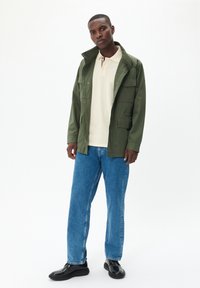 Grüne wasserdichte Jacke mit mehreren Taschen, beige Polohemd, blaue lockere Jeans und schwarze Schuhe vor einem neutralen Hintergrund.
