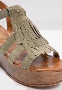 Sandale en suède vert avec un détail en franges, plateforme en bois brun et accents en métal doré. Design à lanières avec un bout ouvert.