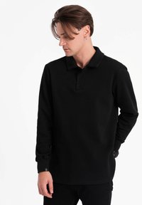 Musta pitkähiha polo-paita puuvilla-sekoitteesta, jossa on perinteinen kaulus, nappilista ja ribatut hihansuut hienovaraisella brändäysyksityiskohdalla.