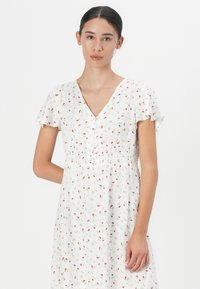 Robe florale blanche ornée de petites roses rouges, avec des manches courtes à volants, un décolleté en V et une fermeture à boutons. Elle possède une taille smockée pour un ajustement décontracté.