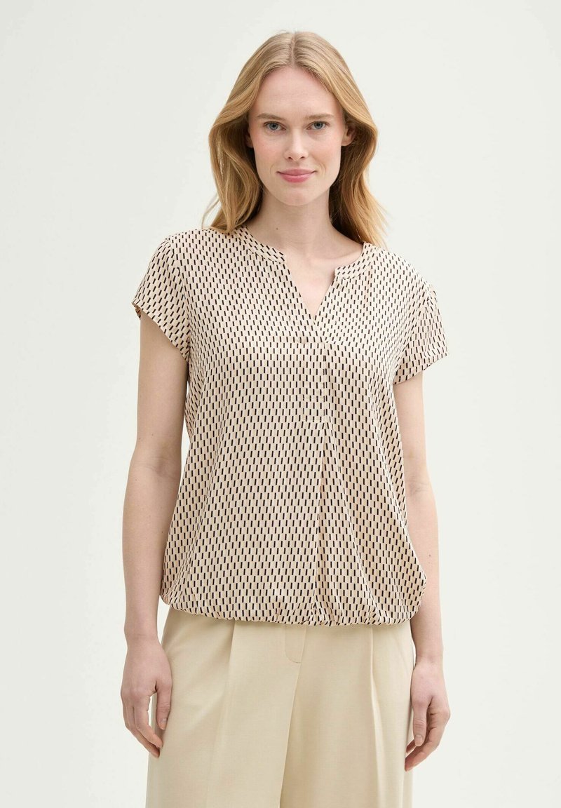 Blouse beige à manches courtes avec un col en V et un motif géométrique noir et blanc, fabriquée en tissu léger avec une coupe ample.