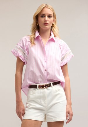 Chemise rose clair à boutons avec des rayures verticales blanches, ourlets des manches rehaussés de dentelle, portée avec un short blanc taille haute et une ceinture marron.