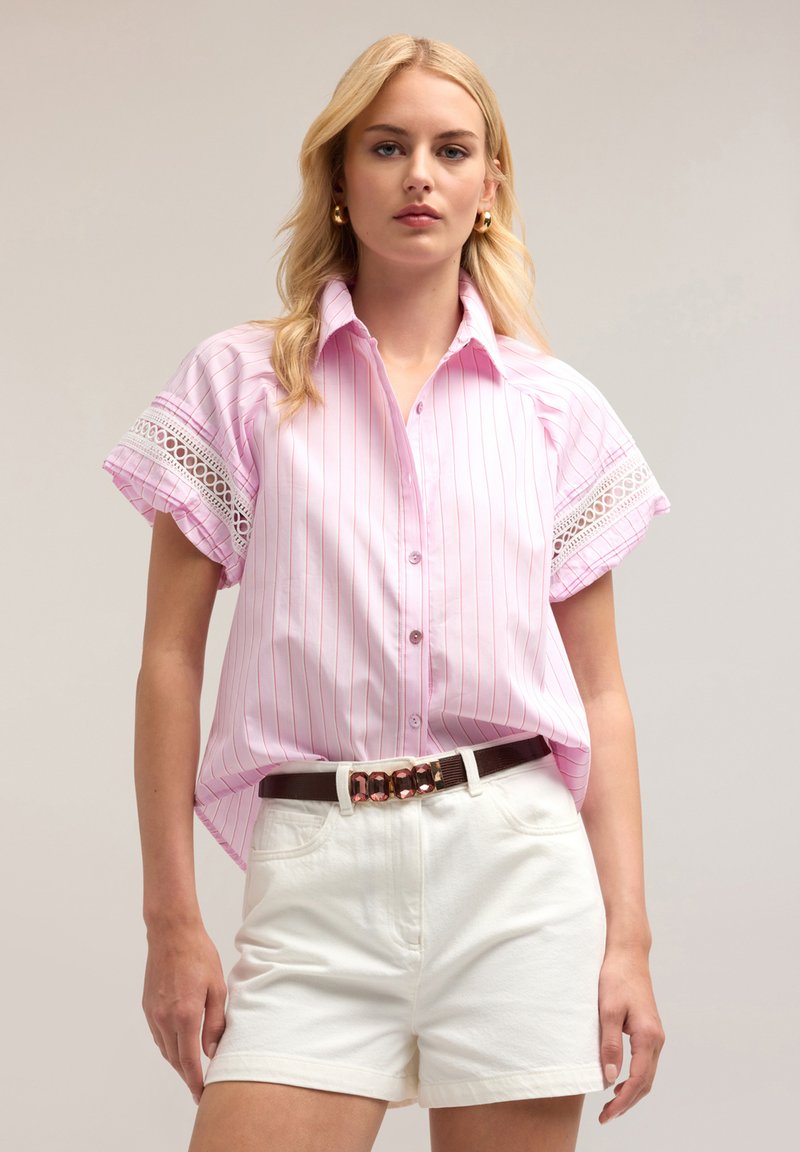 Camicia rosa chiaro con bottoni, strisce bianche verticali, orli delle maniche decorati con pizzo, indossata con pantaloni corti bianchi a vita alta e una cintura marrone.