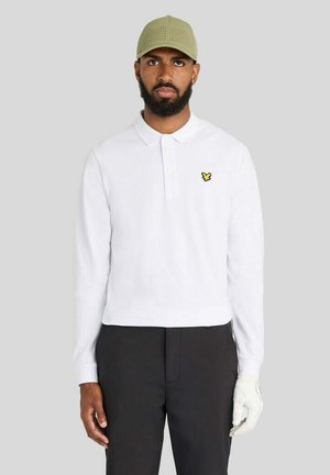 Mann trägt ein weißes langärmeliges Poloshirt, schwarze Hose, beige Kappe und einen weißen Golfhandschuh an der rechten Hand vor einem einfarbigen grauen Hintergrund.