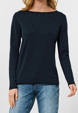 Frau trägt ein schwarzes Langarmshirt mit kleinen silbernen Nieten nahe am Halsausschnitt und hellblaue Jeans, vor einem einfarbigen Hintergrund.