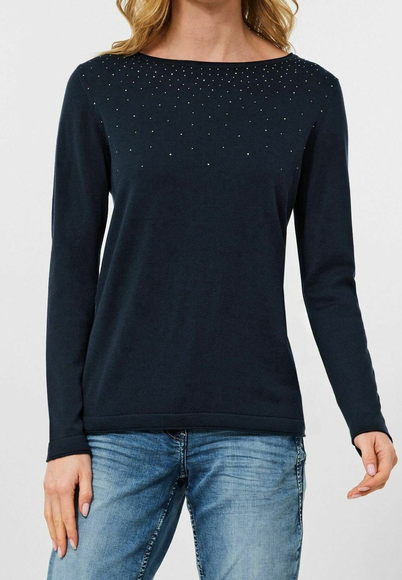 Femme portant un haut noir à manches longues avec de petites clous argentés près de l'encolure et un jean bleu clair, sur un fond uni.