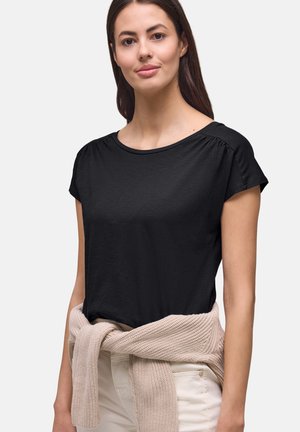 Frau trägt ein schwarzes Kurzarm-Top mit einem beigen Strickpullover, der um die Taille gebunden ist, und beigen Hosen vor weißem Hintergrund.