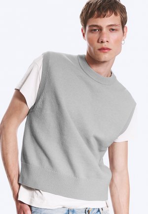 Jeune homme portant un pull sans manches en tricot gris clair sur un t-shirt blanc et un jean bleu clair, debout devant un fond uni.