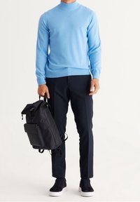 Ljusblå, långärmad tröja med hög krage, tillsammans med mörka byxor, håller en svart ryggsäck med dragkedjestängning och vadderade remmar.