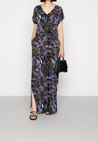 Femme portant une robe maxi noire à fleurs avec des manches courtes, tenant un sac à main noir et portant des sandales noires à lanières.