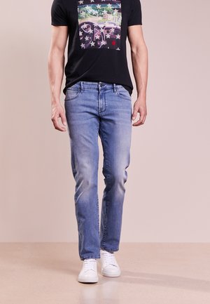 Mand iført lyseblå jeans, hvide sneakers og en sort T-shirt med et farverigt bil- og stjernedesign, står på en neutral baggrund.