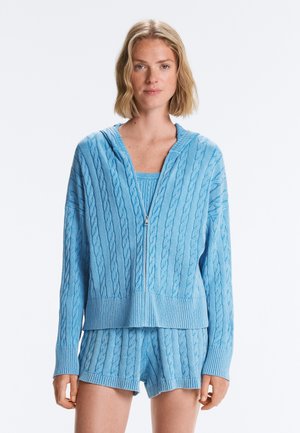 Femme debout portant un sweat à capuche zippé en maille torsadée bleu clair, un short en maille assorti et un débardeur en maille en dessous.