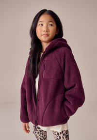 Lys lilla hættetrøje med en tekstureret overflade, lynlås foran og en sidelomme. Sammen med en let cremefarvet top og mønstrede leggings.