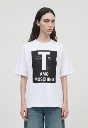 Un mannequin se tient devant un fond uni, vêtu d’un T-shirt blanc orné d’un visuel noir « I AM MOSCHINO » et d’un jean bleu.
