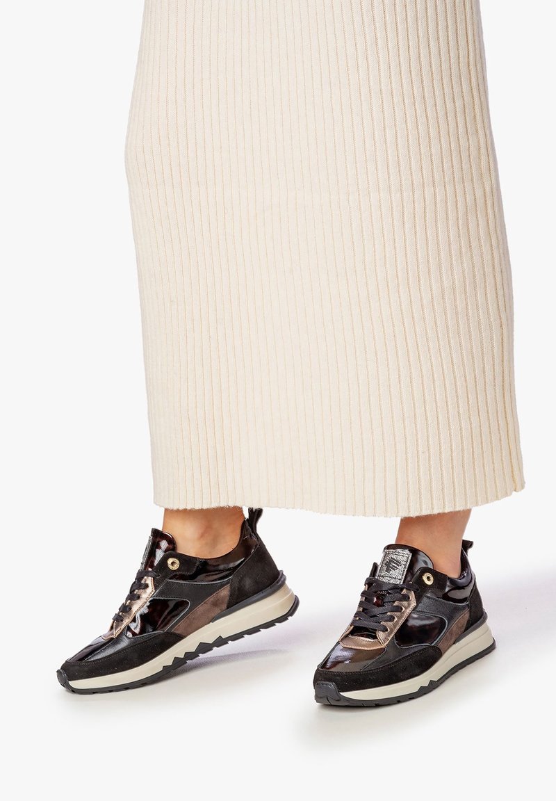 Personne portant des baskets brillantes noires et marron avec des semelles beige, associées à une jupe côtelée crème coupée au niveau du milieu du mollet.