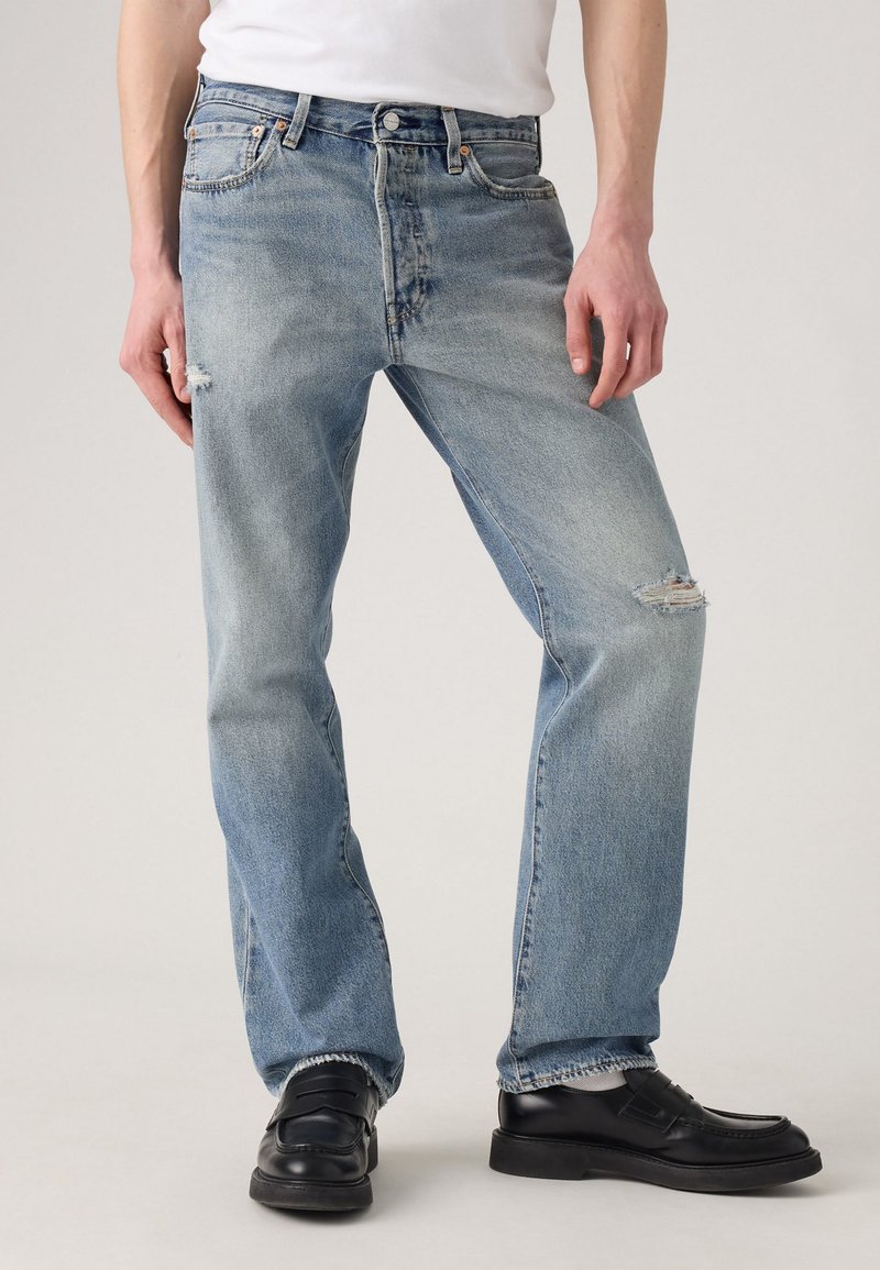 Man wearing light blue ripped jeans, white socks, black leather shoes, and a white shirt shown from waist to feet.