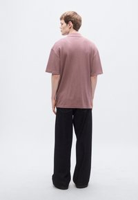 Ροζ υφασμάτινο πουκάμισο με κοντά μανίκια και γιακά, φορεμένο με άνετο μαύρο παντελόνι. Το φόντο είναι απλό και ουδέτερο.