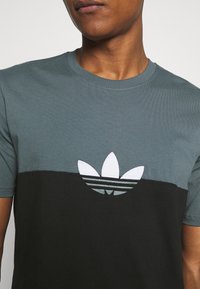 Camiseta de manga corta en verde oscuro y negro con un logo de Adidas bordado en blanco que presenta las tres franjas, cuello redondo y textura acanalada.